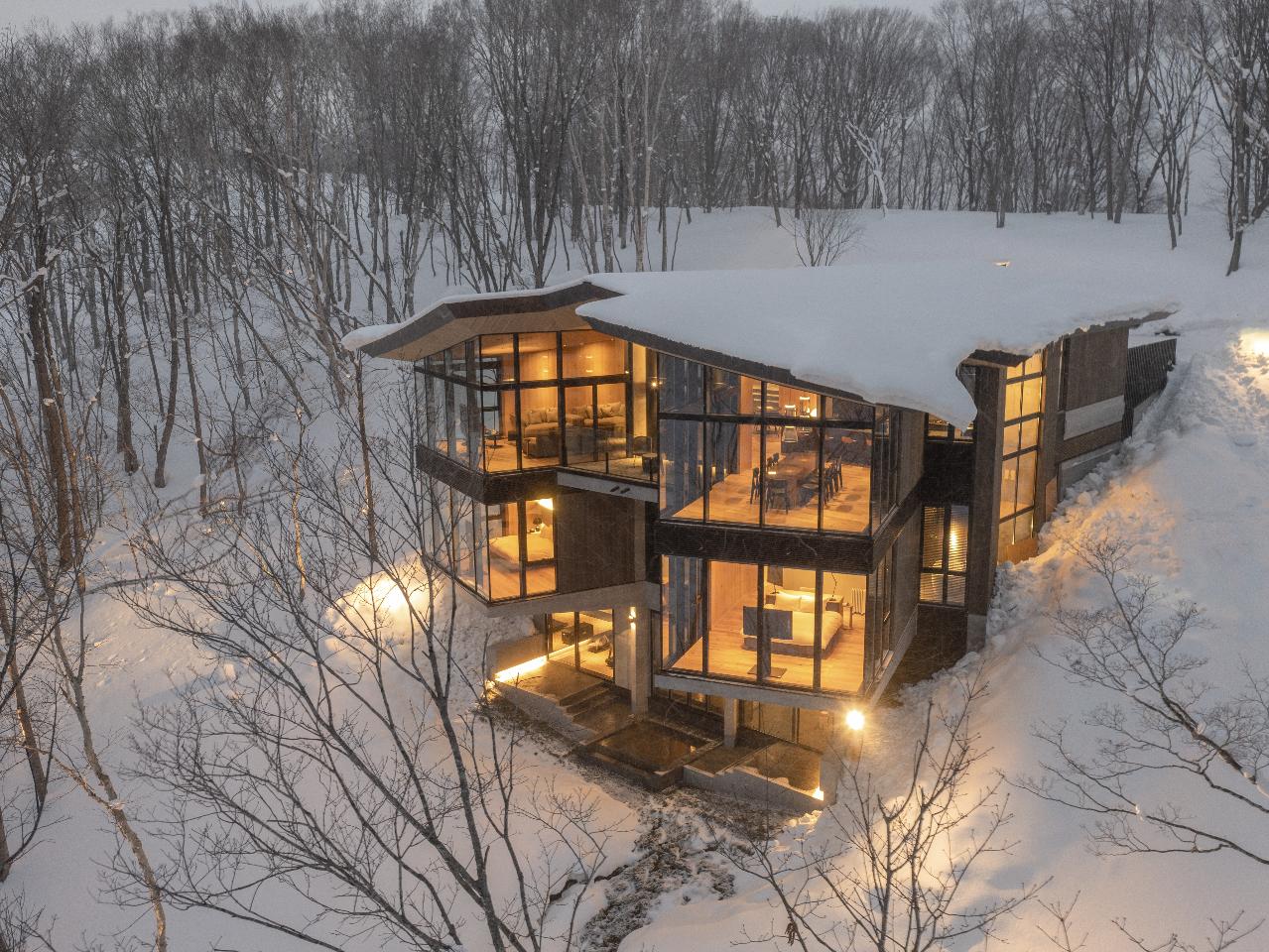 Glass-walled Niseko luxury chalet glowing warmly at dusk amid fresh snow and birch forest