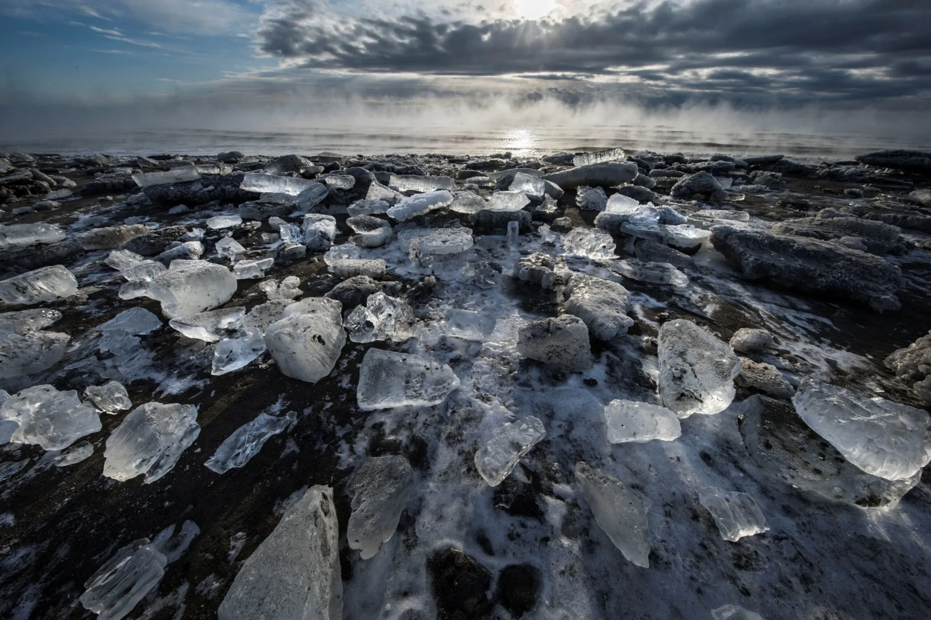 Clear chunks of drift ice scattered across the black shoreline of the Okhotsk Sea under low winter light
