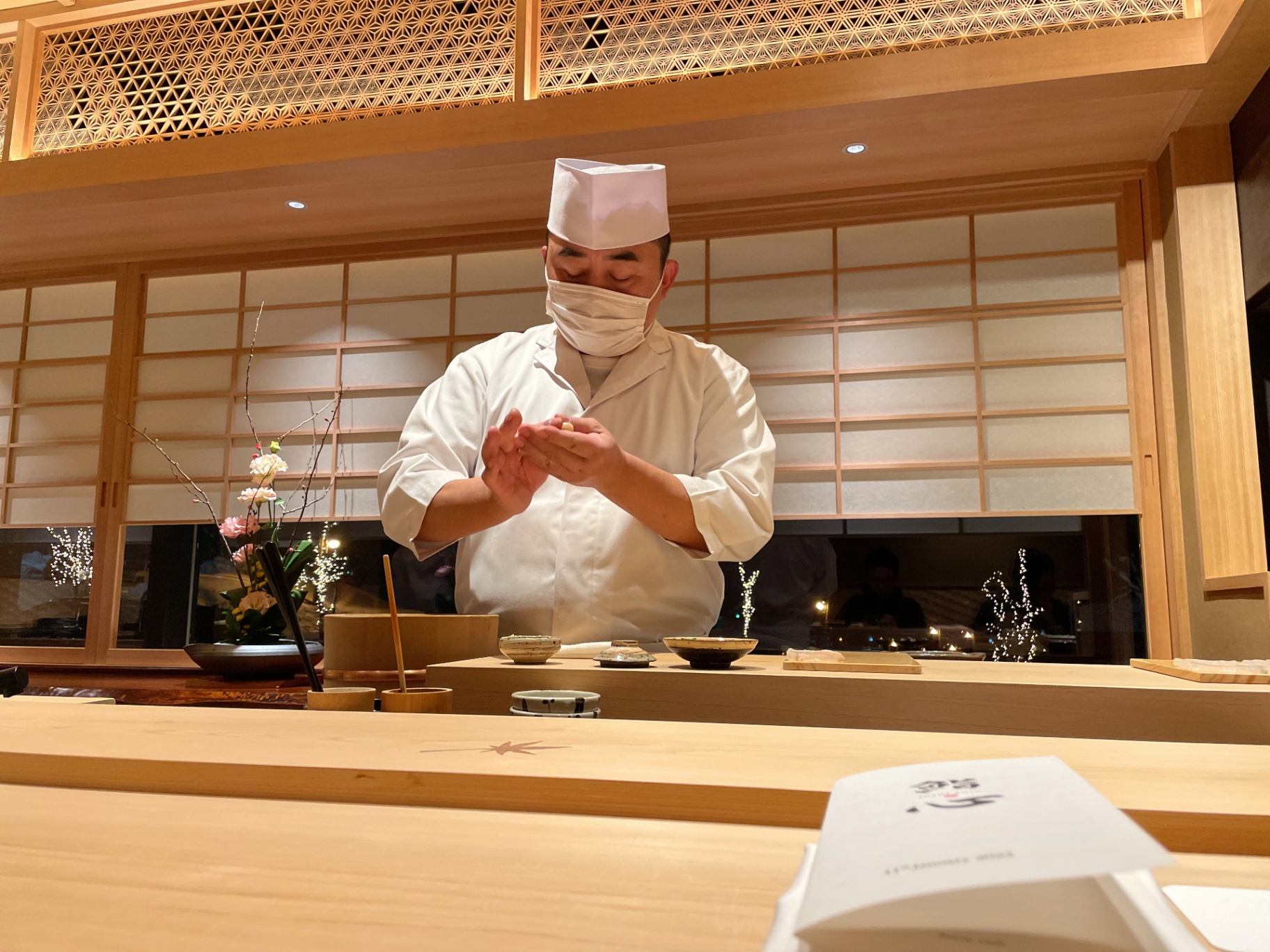 Sushi master forming nigiri at a hinoki counter beneath kumiko lattice