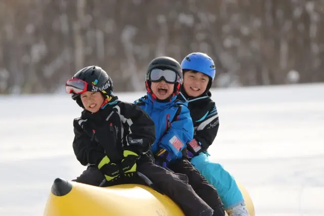 Children in ski helmets laughing on a snow banana boat across a Niseko snowfield