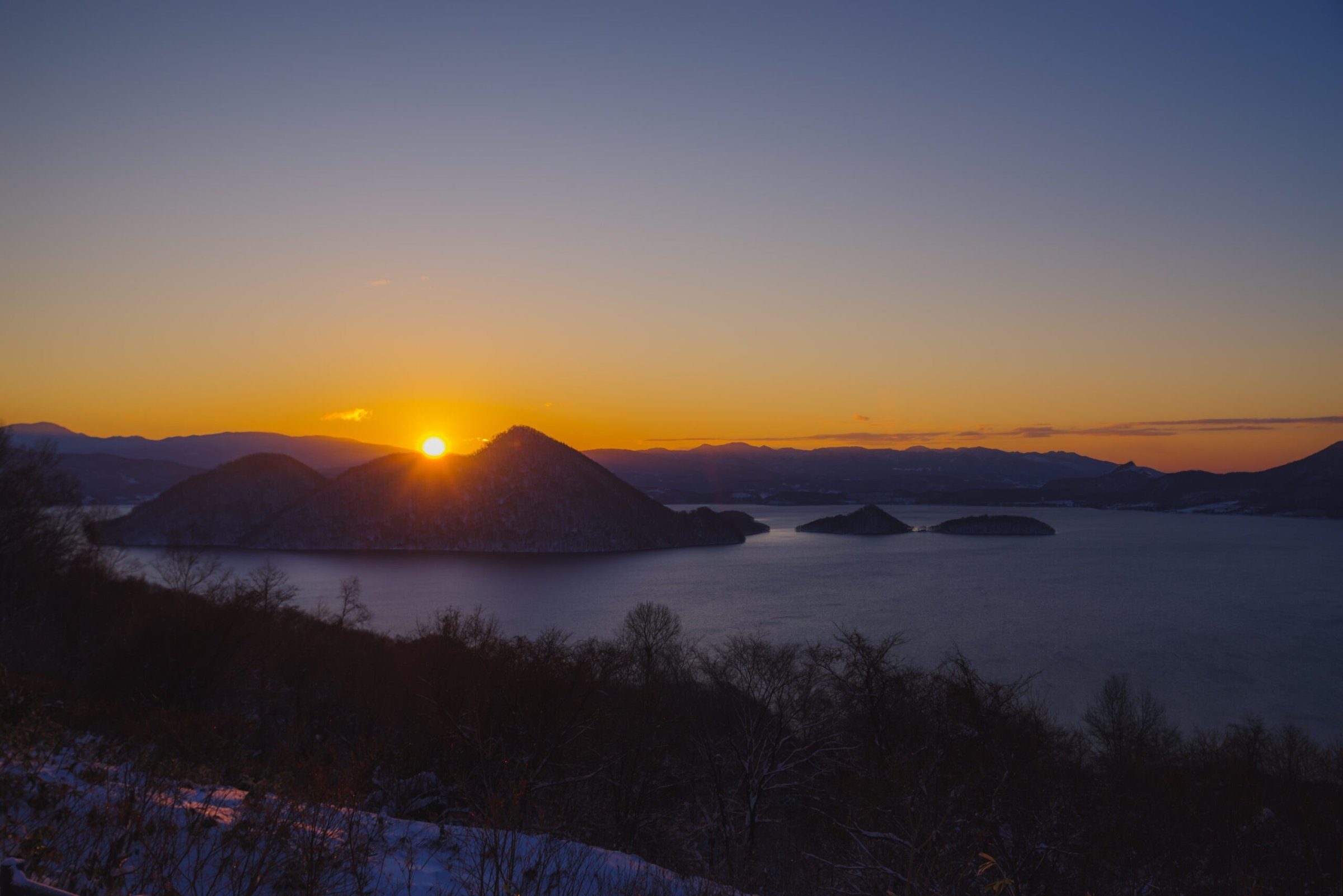 Winter sunrise over Lake Toya and its islands, framed by snowy woodland