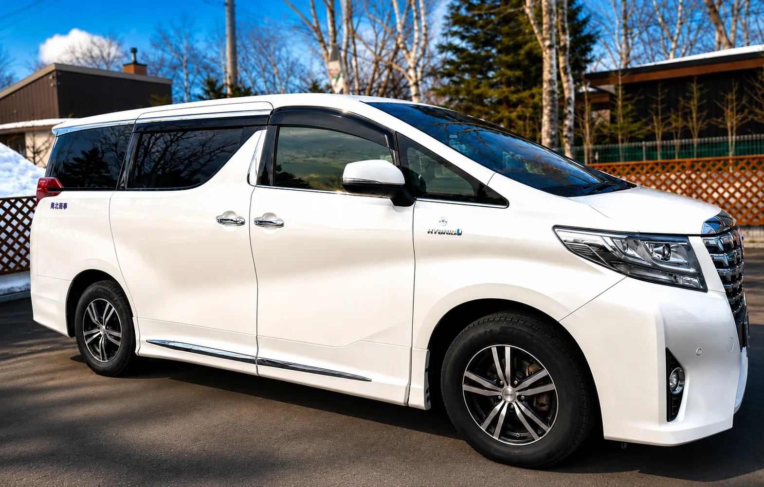 White Toyota Alphard Hybrid private charter vehicle in full side profile on a quiet Hokkaido driveway with birch trees behind